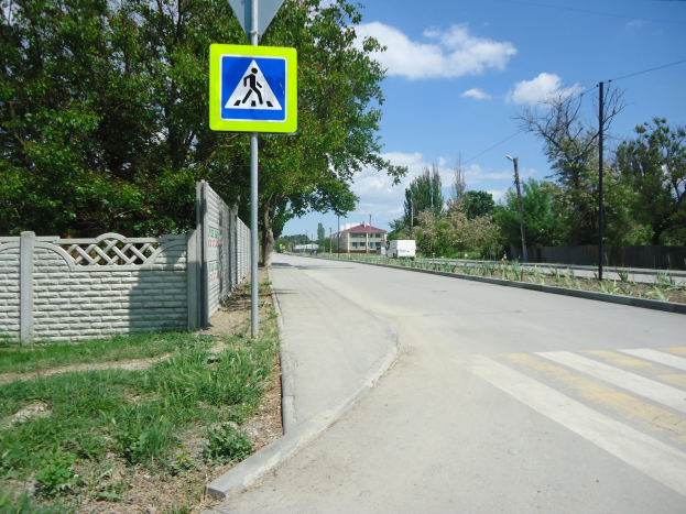 Straße mit einem Fußgängerübergangsschild an der Seite, umgeben von Gras, Pflanzen, einem Zaun, Bäumen, Strommasten mit Drähten, fahrenden Fahrzeugen, entfernten Gebäuden und einem bewölkten Himmel.