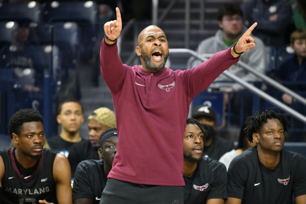 Cheftrainer des Maryland-Männer-Basketballteams in einem marinefarbenen Hemd und schwarzen Shorts, der auf der Seitenlinie einer Basketballhalle gestikuliert, umgeben von sitzenden und stehenden Personen.