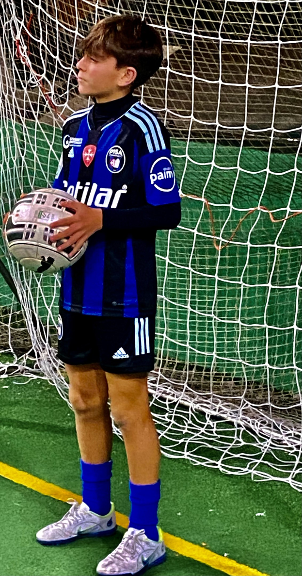 Ein junger Junge steht auf einem Fußballfeld und hält einen Fußball in der Hand, trägt eine Sportuniform und Schuhe und hat ein Netz im Hintergrund.