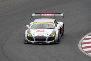 Ein leuchtend rotes und weißes Audi R8 LMS GT3 Rennfahrzeug mit dem Audi-Logo auf der Seite, gefahren von einer Person in einem schwarzen Rennanzug und Helm, fährt auf einer Rennstrecke.
