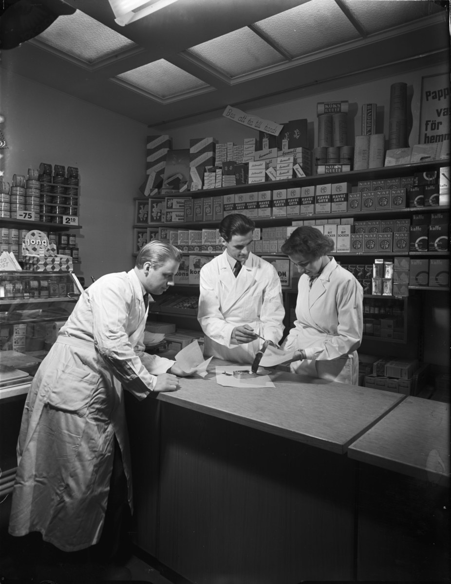 Drei Männer in Laborkitteln an einer Apothekentheke mit Papieren und Gegenständen, vor Regalen mit Artikeln, in Schwarz-Weiß.