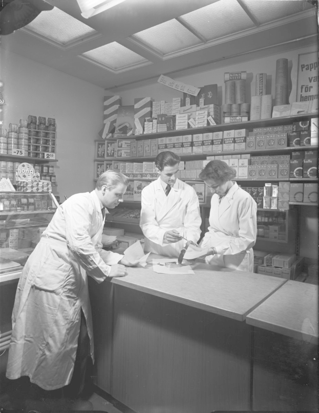 Drei Männer in Laborkitteln an einer Apothekentheke mit Papieren und Gegenständen, vor Regalen mit Artikeln, in Schwarz-Weiß.