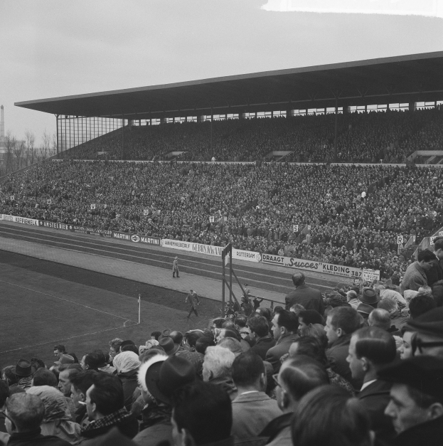 Schwarzes und weißes Foto einer vollen Stadion mit Zuschauern bei einem Fußballspiel, mit Bannern, Pfosten, einem Schuppen, Bäumen, einem Turm und einem bewölkten Himmel.