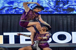 Zwei Performer in violetten Kleidern führen einen akrobatischen Hebemanöver auf der Bühne vor einem Publikum aus, mit einem schwarzen Vorhang und Bildschirm im Hintergrund und Zuschauern unten sichtbar.