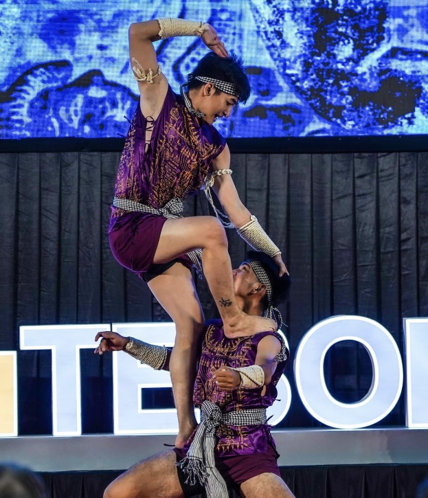Zwei Performer in violetten Kleidern führen einen akrobatischen Hebemanöver auf der Bühne vor einem Publikum aus, mit einem schwarzen Vorhang und Bildschirm im Hintergrund und Zuschauern unten sichtbar.