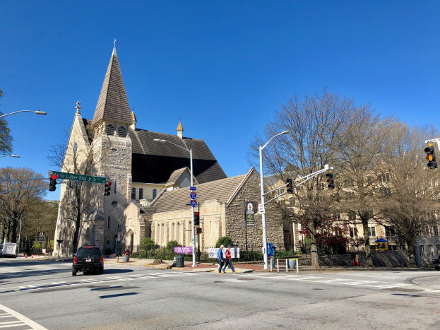 Große Kirche mit Turm an der Straßenecke, identifiziert als St. Lukaskirche, umgeben von Gebäuden, Straßenmöbeln, Fahrzeugen, Fußgängern, Grünflächen und einem klaren blauen Himmel.