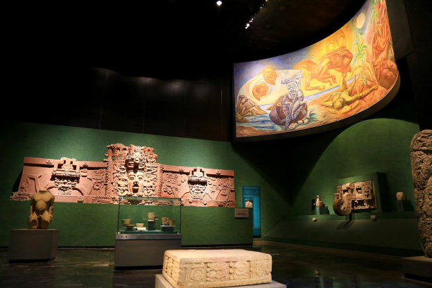 Ausstellungsraum im Anthropologischen Museum von Mexiko-Stadt mit verschiedenen Artefakten, darunter Skulpturen, Tischobjekte, eine Texttafel, eine Wandmalerei und Deckenbeleuchtung.