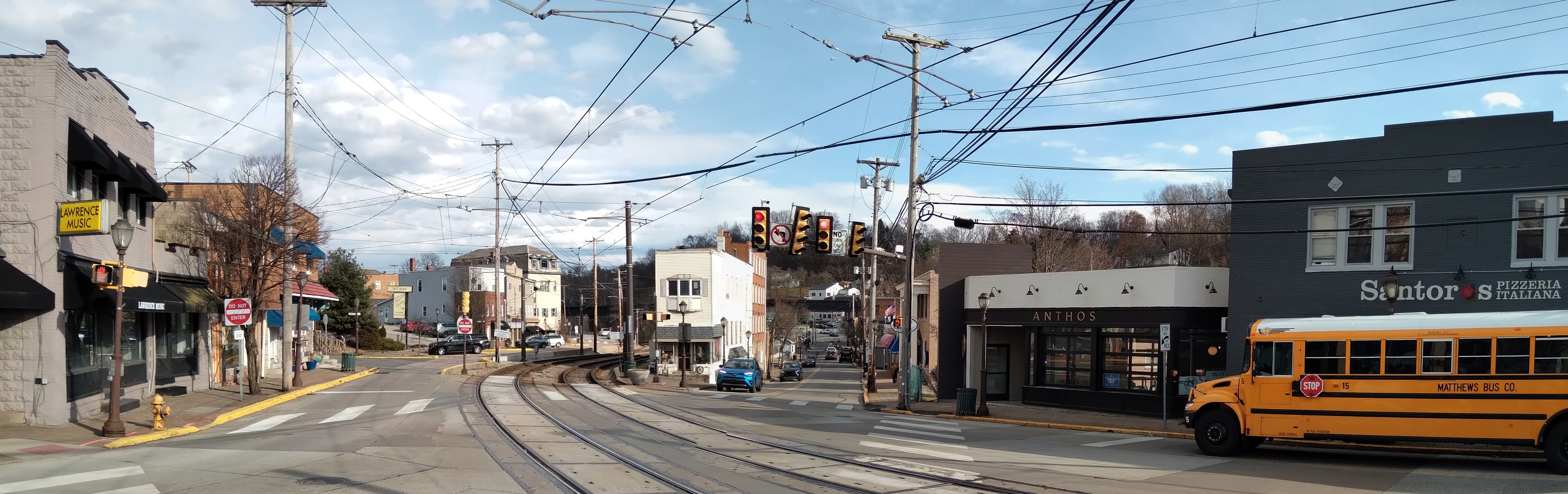 Ein gelber Schulbus fährt auf einer Straße mit hohen Gebäuden, Strommasten, Ampeln, Schildern, Bäumen und einem bewölkten Himmel.