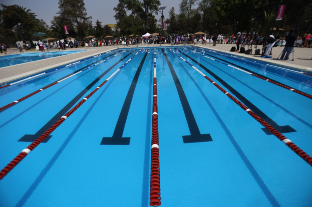 Gro├čes Schwimmbad mit Bahnentrennungen, umgeben von Menschen mit Taschen und Gegenst├Ąnden, mit Zelten, Transparenten, Masten, einer Anzeigetafel und einem bew├Âlktem Himmel im Hintergrund.