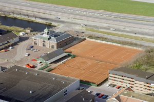 Eine Luftaufnahme eines Tennisplatzes in einer Stadt, umgeben von Gebäuden, Fahrzeugen, Straßen, Gras, Bäumen und Wasser.