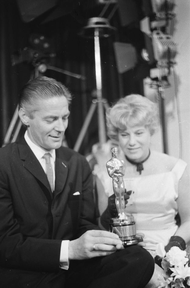 Ein Mann in einem schwarzen Blazer, einem weißen Hemd und einer Krawatte hölt eine Oscar-Statue, waehrend er neben einer Frau in Abendgarderobe sitzt, die einen Blumenstrau├č hölt, mit weicher Beleuchtung im Hintergrund.