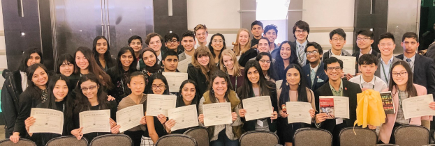 Gruppe von Menschen mit Urkunden bei einer Abschlussfeier mit Stühlen und einer Wand im Hintergrund.