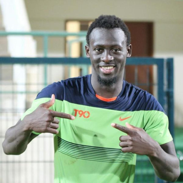 Ein Mann in einem grün-blauen Hemd, der grinsend in die Kamera zeigt und im Hintergrund eine Brüstung, eine Wand und eine Tür zu sehen sind.