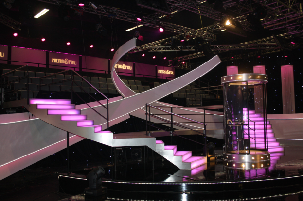 Bühne mit Wendeltreppe, Geländern, Stufen, Beleuchtungsequipment, vertikalen Stangen, Texttafeln, Säulen und anderen Requisiten vor dunklem Hintergrund.