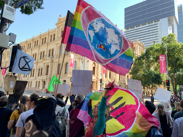 Eine Gruppe von Menschen mit Fahnen und Schildern vor einem Gebäude, umgeben von Bäumen, Laternenmasten, Verkehrszeichen und Tafeln mit Text, bei einer Schwulen- und Lesbenparade in Melbourne.