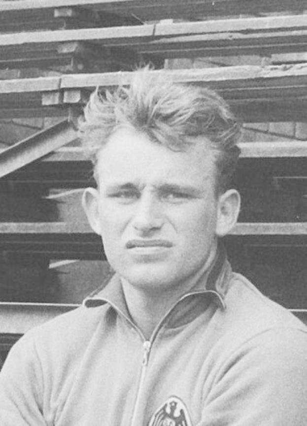 Schwarzes und weißes Foto eines Mannes in einer Jacke, der vor einem Haufen Holzplanken steht, der angeblich ein legendärer deutscher Fußballspieler ist.