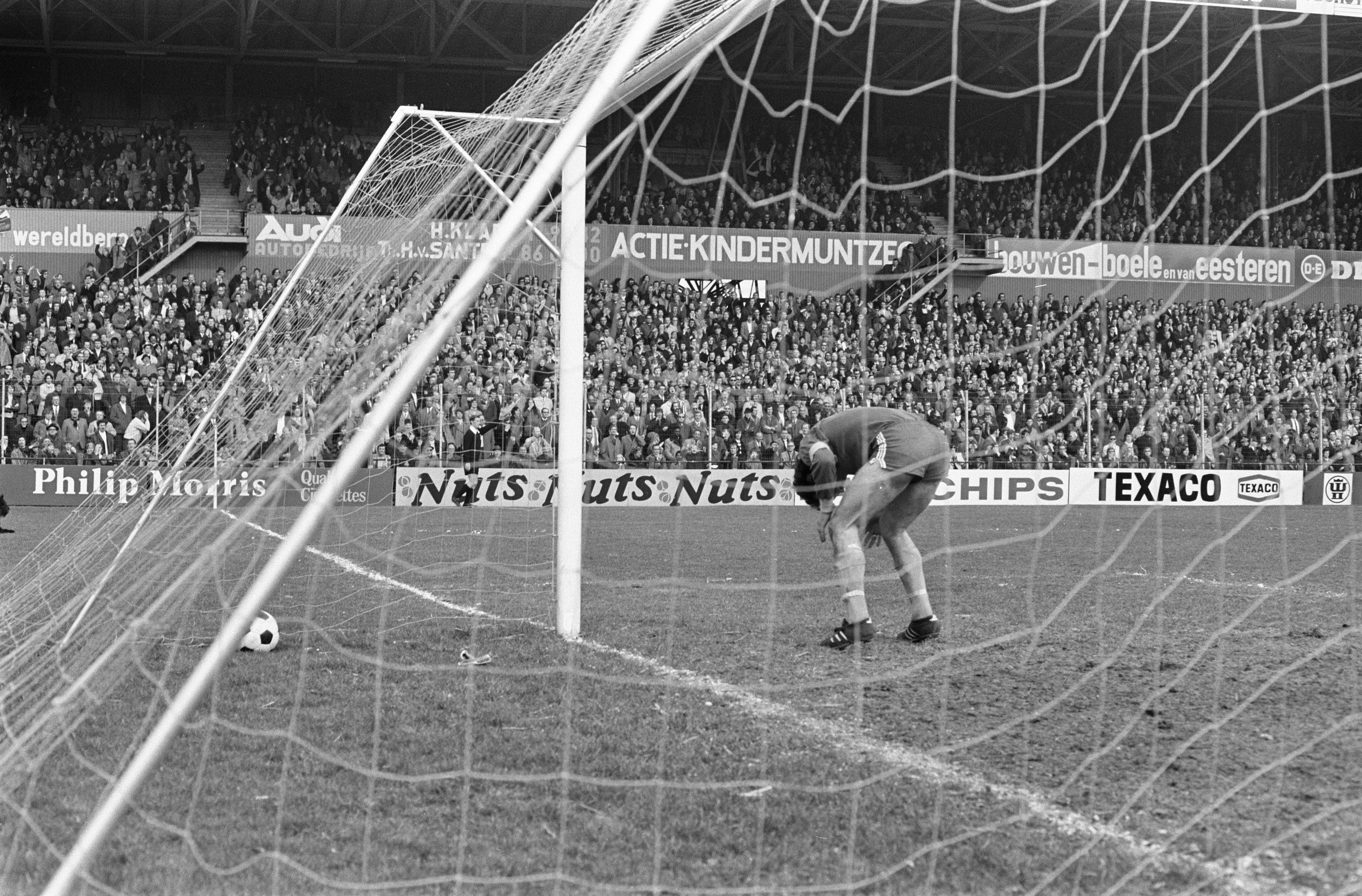 Schwarzes und weißes Foto eines Fußballtorwarts, der einen Ball in der Nähe des Pfostens abwehrt, mit Zuschauern und Bannern im Hintergrund.