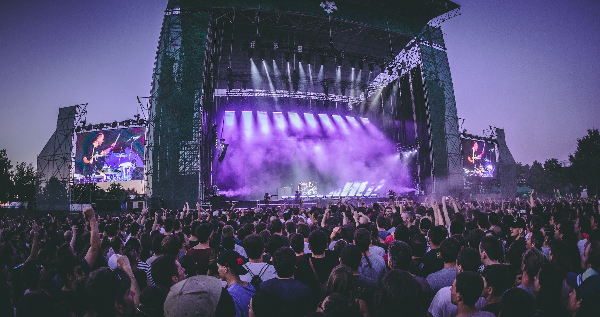 Eine große Menschenmenge auf einem Musikfestival schaut Musikern auf einer hell erleuchteten Bühne mit Instrumenten und Bildschirmen zu, die vor einem Gebäude, Bäumen und einem klaren blauen Himmel aufgebaut sind.