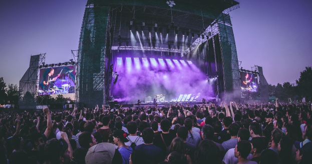Eine große Menschenmenge auf einem Musikfestival schaut Musikern auf einer hell erleuchteten Bühne mit Instrumenten und Bildschirmen zu, die vor einem Gebäude, Bäumen und einem klaren blauen Himmel aufgebaut sind.