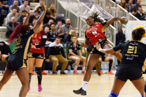 Eine Gruppe von Frauen spielt Volleyball auf einem Feld mit einem Ball in der Mitte, während Zuschauer auf Stühlen und Geländern im Hintergrund sitzen und Flaschen auf dem Boden liegen.
