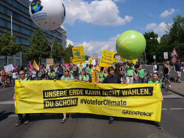 Eine Gruppe von Menschen geht eine Straße entlang und hält ein gelbes "Wähle für das Klima"-Plakat mit Luftballons, Schildern, Fahrrädern, Bäumen, Laternenmasten und Gebäuden im Hintergrund unter einem klaren blauen Himmel.