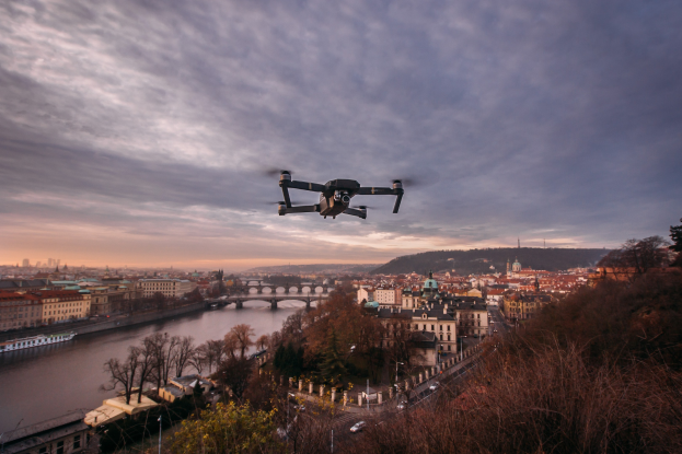 Drohne fliegt über einer Stadt mit einem Fluss, Gebäuden, Bäumen, Fahrzeugen und einer Brücke im Hintergrund unter einem sichtbaren Himmel.