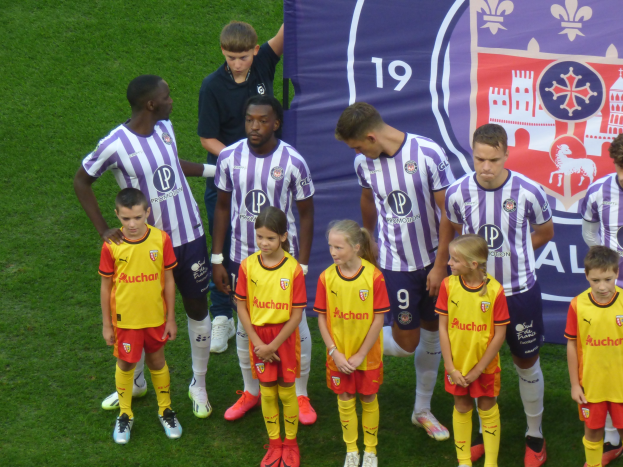 Eine Gruppe junger Fußballspieler steht auf einem saftig grünen Feld unter einem klaren blauen Himmel, mit einem "Ligue 1"-Schild im Hintergrund.
