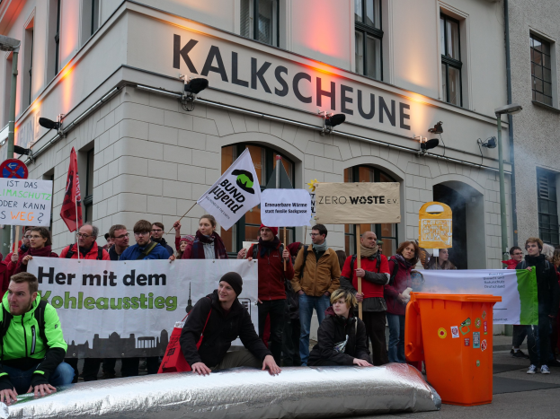 Eine Gruppe von Menschen mit Protestschildern vor einem Gebäude, mit zwei Personen im Vordergrund und einem Müllcontainer auf der rechten Seite.