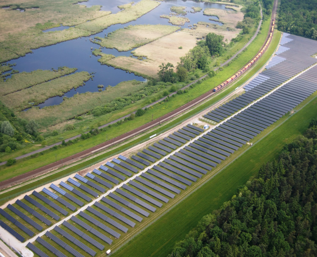 Luftaufnahme einer Solarstromanlage mit Panelen, umgeben von Bäumen, Gras, Wasser und einer nahen Bahnlinie.