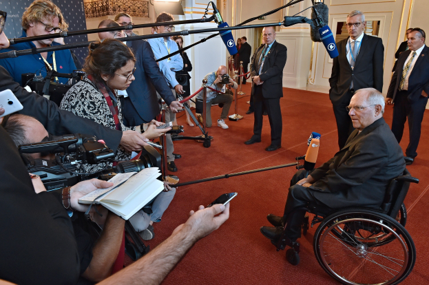 Ein Mann im Rollstuhl wird von Reportern umgeben, die Kameras, Handys und Mikrofone mit Ständern halten, mit einem Banner, Scheinwerfern und einer Wand im Hintergrund.