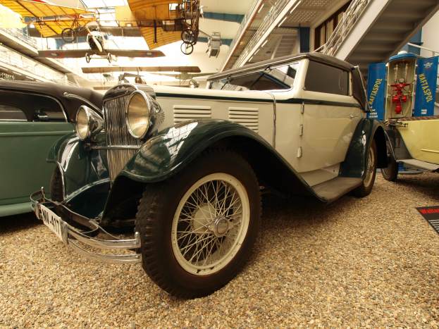 Ein Oldtimer-Auto in einem Museum zwischen anderen Fahrzeugen ausgestellt, mit informativem Text auf einer Tafel rechts und Flugzeugen, die von der Decke hängen.