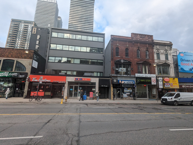 Stadtstraße mit hohen Gebäuden, fahrenden Fahrzeugen, Fußgängern, einem Fahrrad, einem Feuerhydranten und Werbetafeln für Vermietungen unter einem bewölkten Himmel.