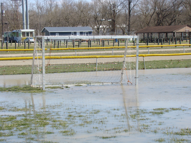 Ein Fußballtor steht in der Mitte eines überfluteten Feldes, umgeben von Gras und Wasser, vor dem Hintergrund von Hütten, Pfählen, Bäumen, Fahrzeugen und einem klaren blauen Himmel.