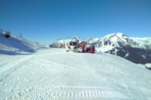 Ein Hubschrauber steht auf einem schneebedeckten Berggipfel, umgeben von ein paar Menschen, unter einem klaren Himmel.