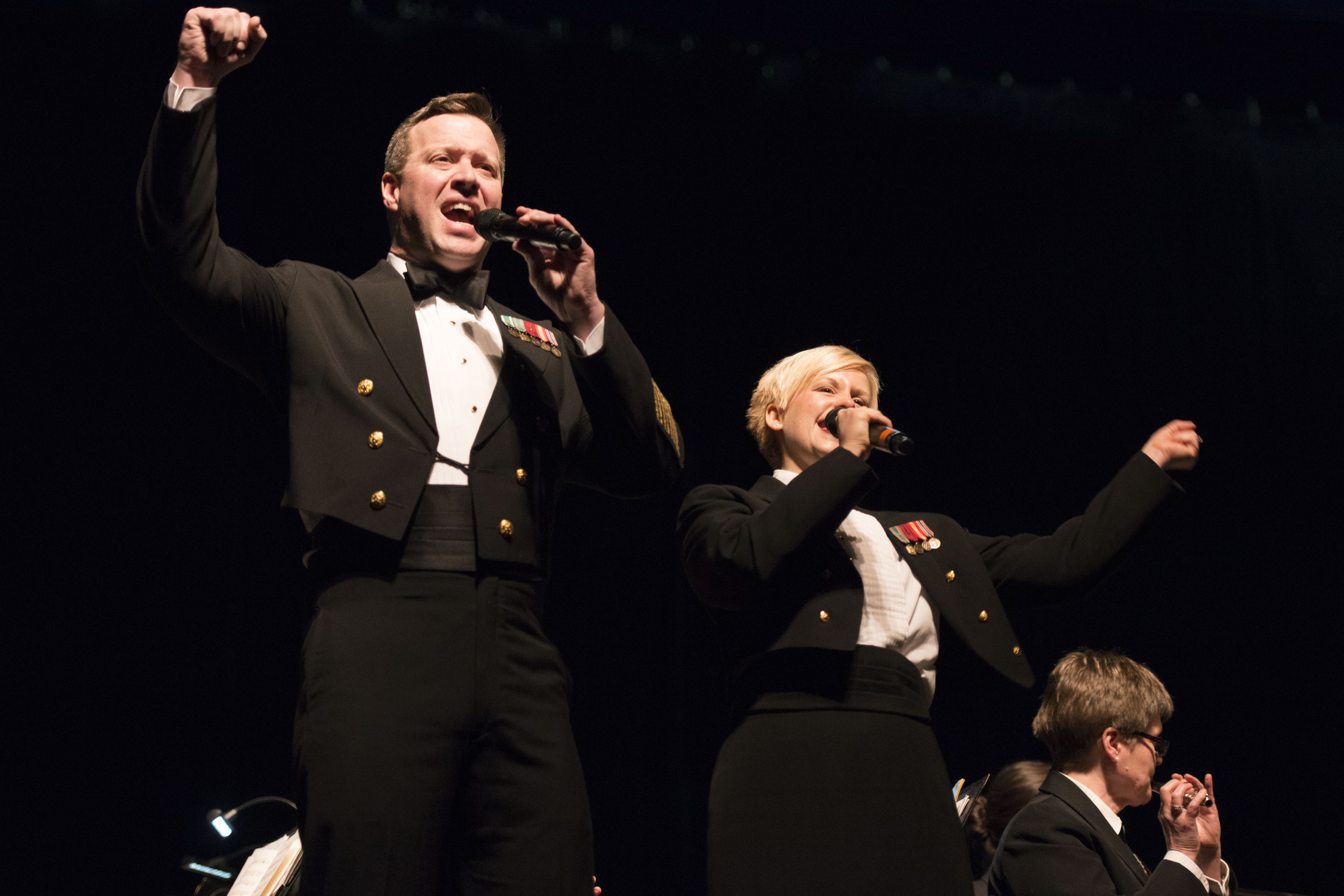 Zwei Militärangehörige in Uniform auf der Bühne, einer hält ein Mikrofon und der andere trägt eine schwarze Jacke und ein weißes Hemd, während das Publikum auf Stühlen gegen einen schwarzen Hintergrund sitzt.