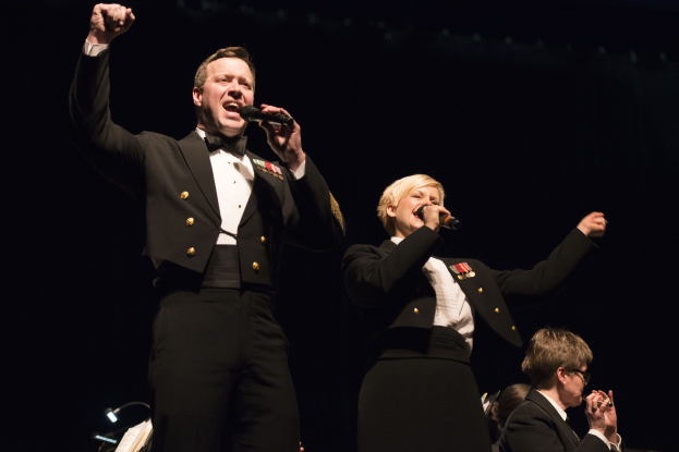 Zwei Militärangehörige in Uniform auf der Bühne, einer hält ein Mikrofon und der andere trägt eine schwarze Jacke und ein weißes Hemd, während das Publikum auf Stühlen gegen einen schwarzen Hintergrund sitzt.