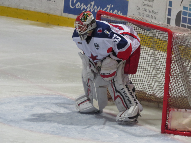 Ein Eishockeytorwart in voller Ausrüstung steht im Netz auf einem Eisstadion, mit einer Wand mit Text im Hintergrund.
