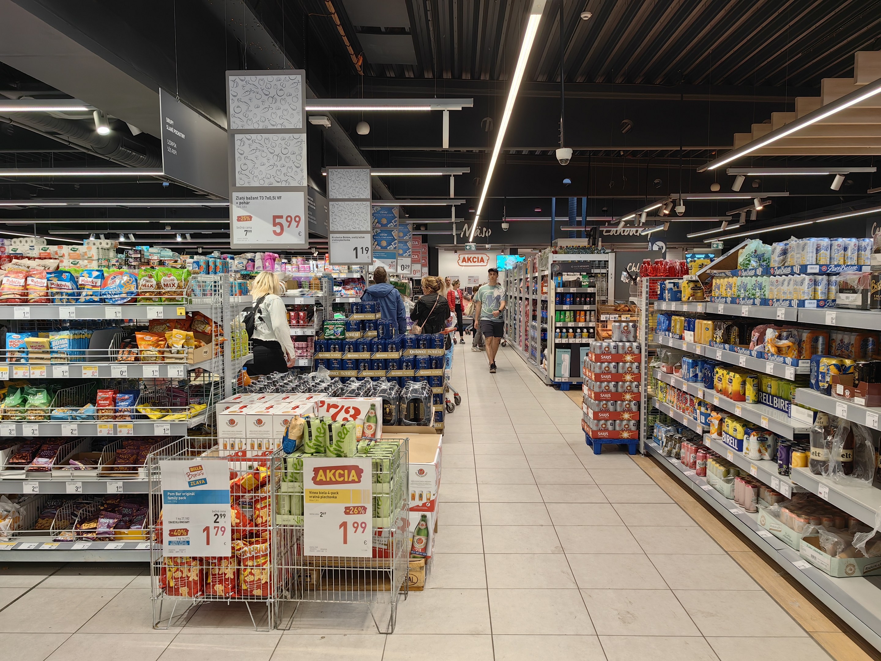 Innensicht eines Supermarkts mit Einkaufenden, Lebensmittelregalen, Einkaufswagen mit Preisschildern, Deckenleuchten, Informationsschildern und einem sichtbaren Boden.