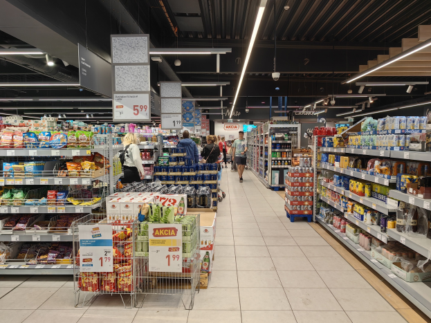 Innensicht eines Supermarkts mit Einkaufenden, Lebensmittelregalen, Einkaufswagen mit Preisschildern, Deckenleuchten, Informationsschildern und einem sichtbaren Boden.