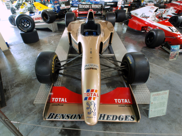 Rennwagen in einem Motorsportmuseum ausgestellt, getrennt durch einen Metallzaun mit Informationsschilder im Hintergrund.