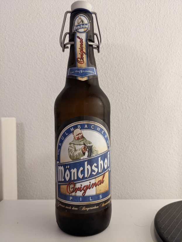 Eine Flasche Münchner Bier mit einem Aufkleber auf einem weißen Tisch, mit einem schwarzen Objekt rechts daneben und einer weißen Wand im Hintergrund.