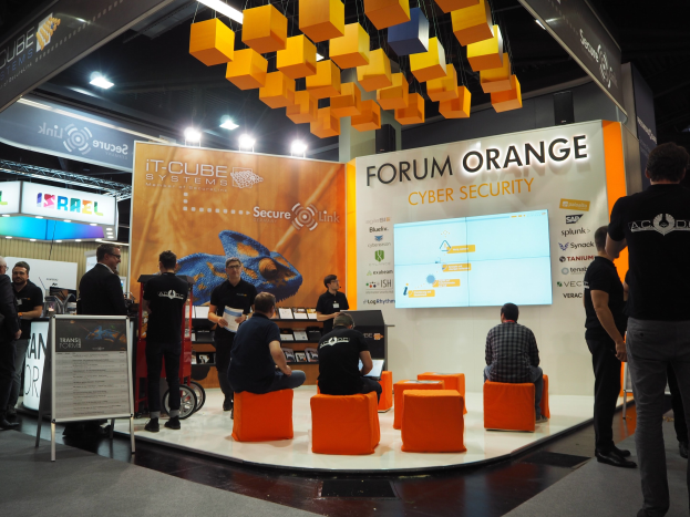 Eine Gruppe von Menschen an der IT-Cube Systems Forum Orange Cyber Security Theke, einige sitzen und andere stehen, in der Nähe einer Tafel, eines Bildschirms, von Regalen, Schilden, Deckenleuchten und Lampen.