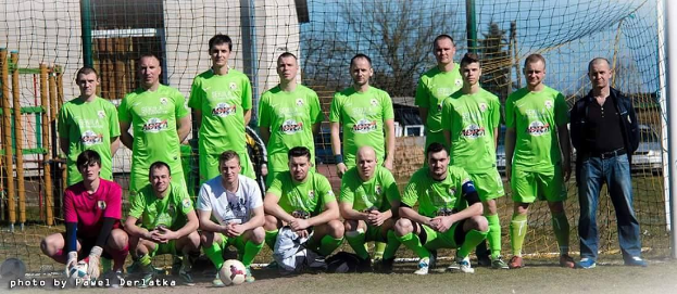 Gruppe von Männer in grünnen T-Shirts auf einem Fussballfeld mit einem Ball und einem Tor, Bäume und Gebäude im Hintergrund.