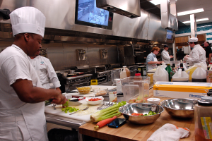 Eine Gruppe von Köchen in einer Küche, die Essen auf einem Schneidbrett zubereiten, mit einem Koch im Vordergrund in einer weißen Schürze und Mütze, der ein Messer hält, umgeben von Utensilien, Schüsseln, Gemüse, einem Handy, Flaschen und anderen Gegenständen, mit einem Fernseher und Deckenlampen im Hintergrund.
