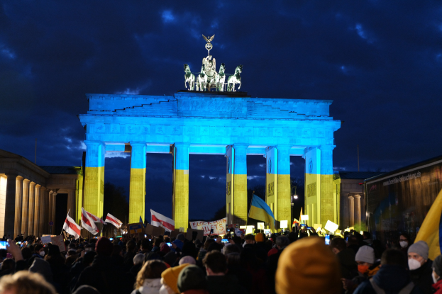 Eine Menge steht vor dem Reichstagsgebäude in Berlin, mit Fahnen und Schildern, einem Banner auf der rechten Seite und bewölktem Himmel.