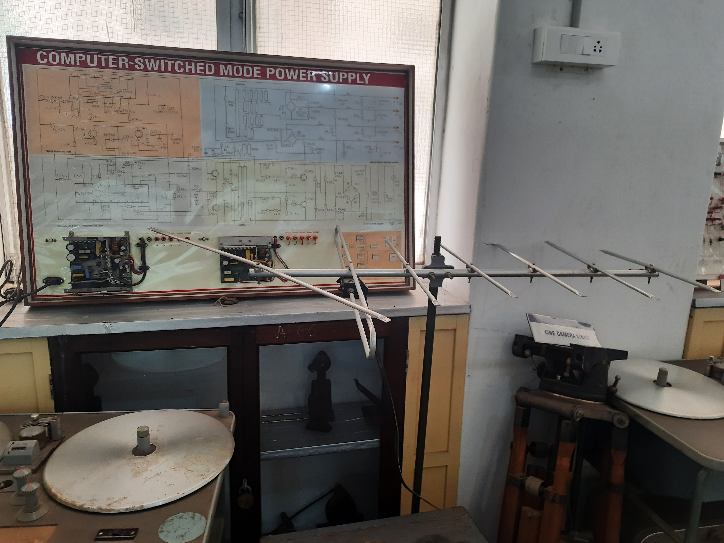 Ein unordentlicher Raum mit verschiedenen Elektronikgeräten, darunter eine beschriftete Platine auf einem Tisch, Metallstangen, eine wandmontierte Schalttafel, ein Fenster und Gegenstände in Schränken, die auf eine computergeschaltete Modussteuerung hindeuten.