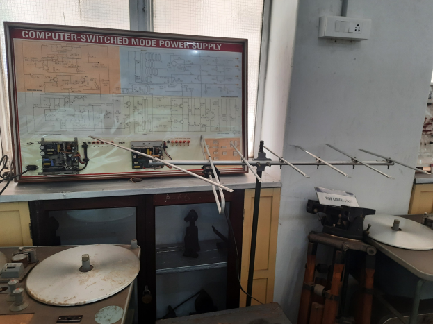 Ein unordentlicher Raum mit verschiedenen Elektronikgeräten, darunter eine beschriftete Platine auf einem Tisch, Metallstangen, eine wandmontierte Schalttafel, ein Fenster und Gegenstände in Schränken, die auf eine computergeschaltete Modussteuerung hindeuten.