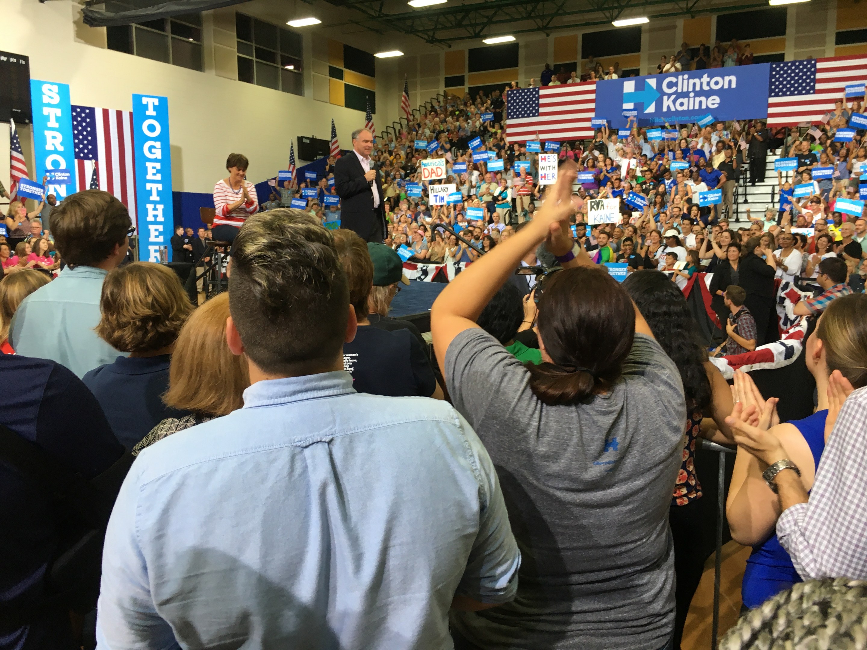 Eine große Menge klatscht und jubelt vor einer Bühne, auf der ein Mann in ein Mikrofon spricht, während eine Frau neben ihm steht; Fahnen, Banner und ein Lautsprecher sind auf dem Bühnenhintergrund zu sehen.