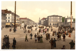 Ein altes Foto einer belebten Münchner Straße in den frühen 1900er Jahren, mit Menschen, die spazieren gehen, einige mit Schirmen, und einem Pferdewagen im Vordergrund, Gebäuden, Bäumen und Polen im Hintergrund und Text am unteren Rand des Bildes.