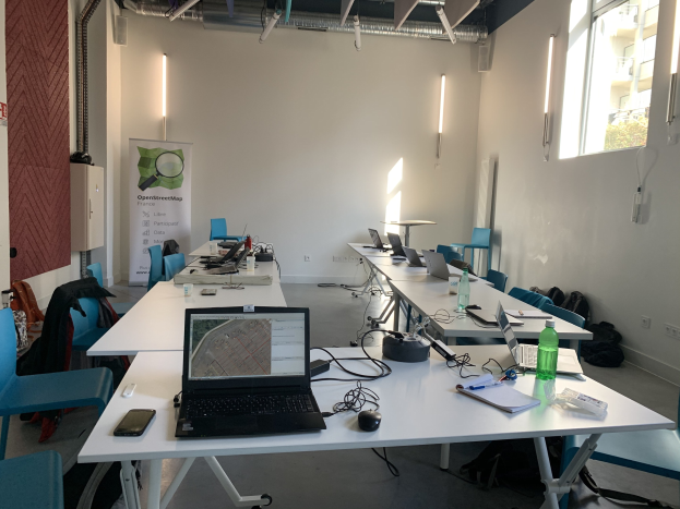 Ein Raum mit mehreren Tischen und Stühlen, die für einen openstreetmap-Workshop in Paris eingerichtet sind, wobei jeder Tisch Laptops, Handys, Bücher, Papiere und andere Gegenstände enthält, sowie Taschen auf dem Boden, eine Wandtafel und Deckenleuchten sowie Fenster.